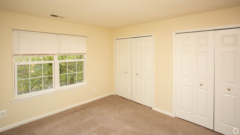 Empty room with beige carpet, cream walls, two closets with white doors, and a window with blinds.