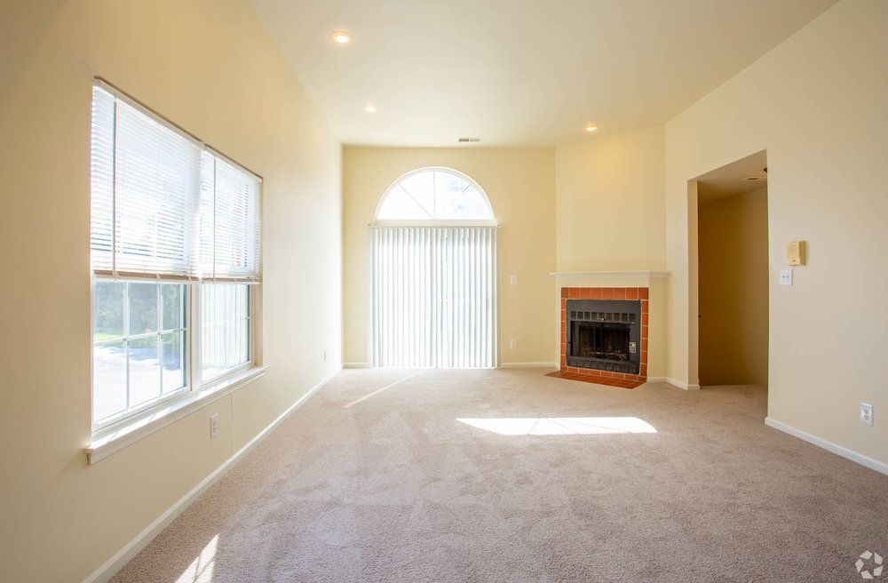 Bright, empty living room with carpet, large windows, fireplace, and sliding glass doors with vertical blinds.