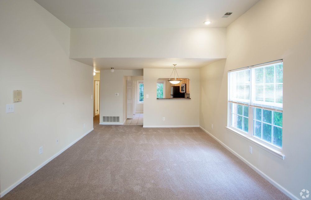 Bright, empty living room with beige carpet, cream walls, large window, and a view into the kitchen area.