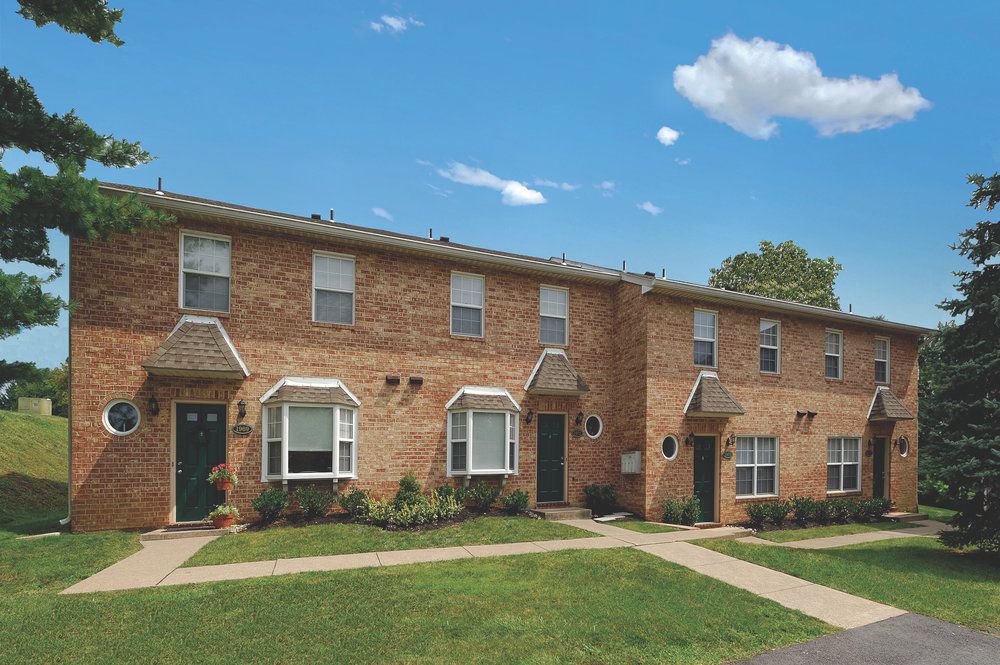 Two connected brick townhouses with green doors, white trim, and a grassy lawn on a sunny day.